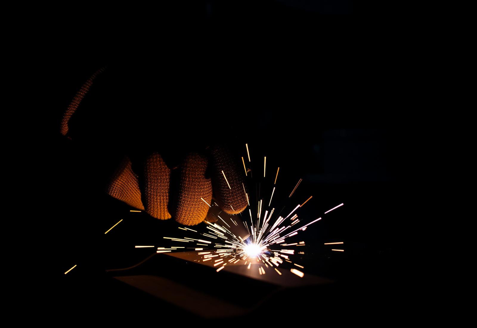 A fabricator's gloved hand welding steel, sparks lit against a black workshop